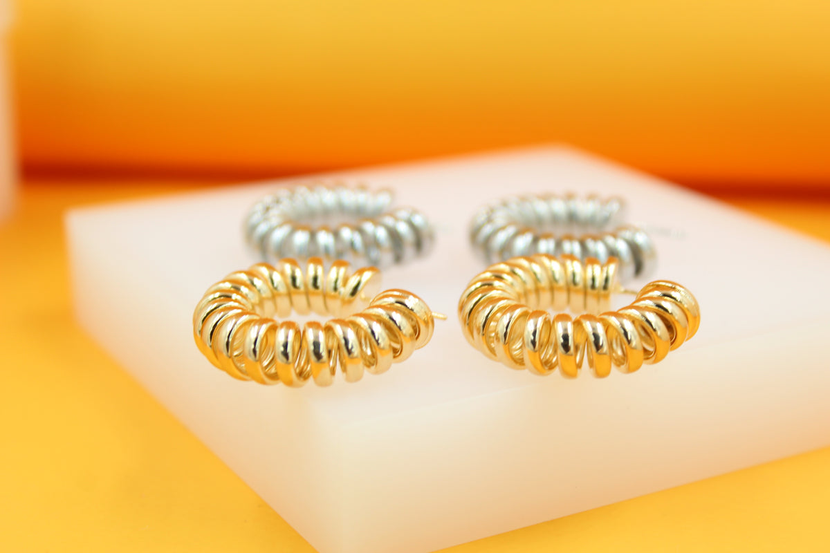 Two pairs of polished gold and silver spiral hoop earrings rest on a white display block against an orange backdrop.