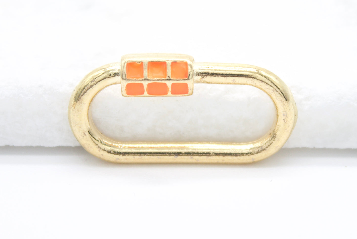 A gold oval carabiner jewelry clasp features vibrant orange enamel details against a bright white background.