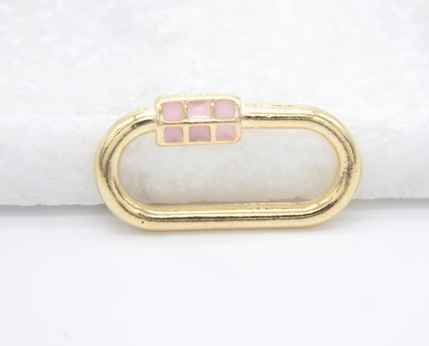 A gold oval lock charm features a pink enamel grid design resting on a white surface.