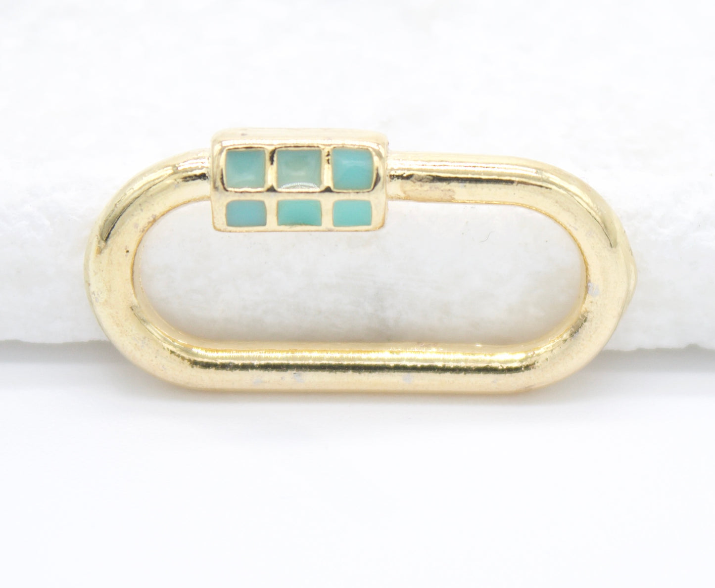 A gold oval carabiner lock charm features a turquoise-inlaid closure mechanism resting on a white surface.