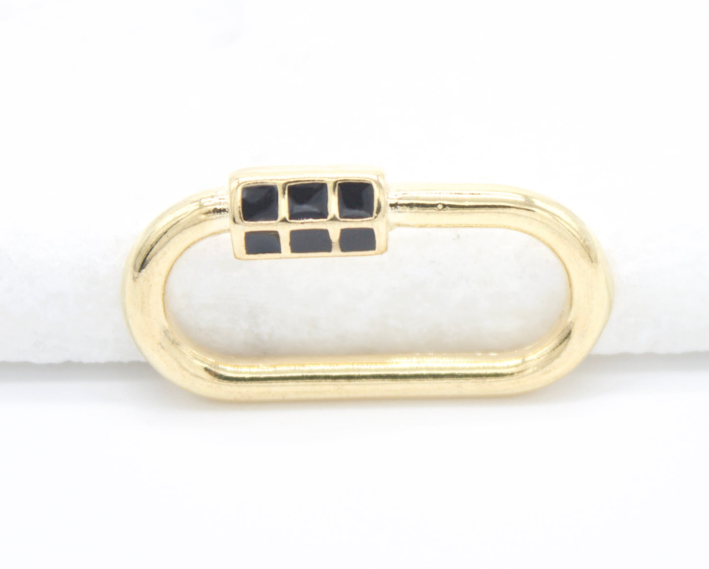 A gold oval charm connector features a black enamel grid pattern on its clasp against a white background.