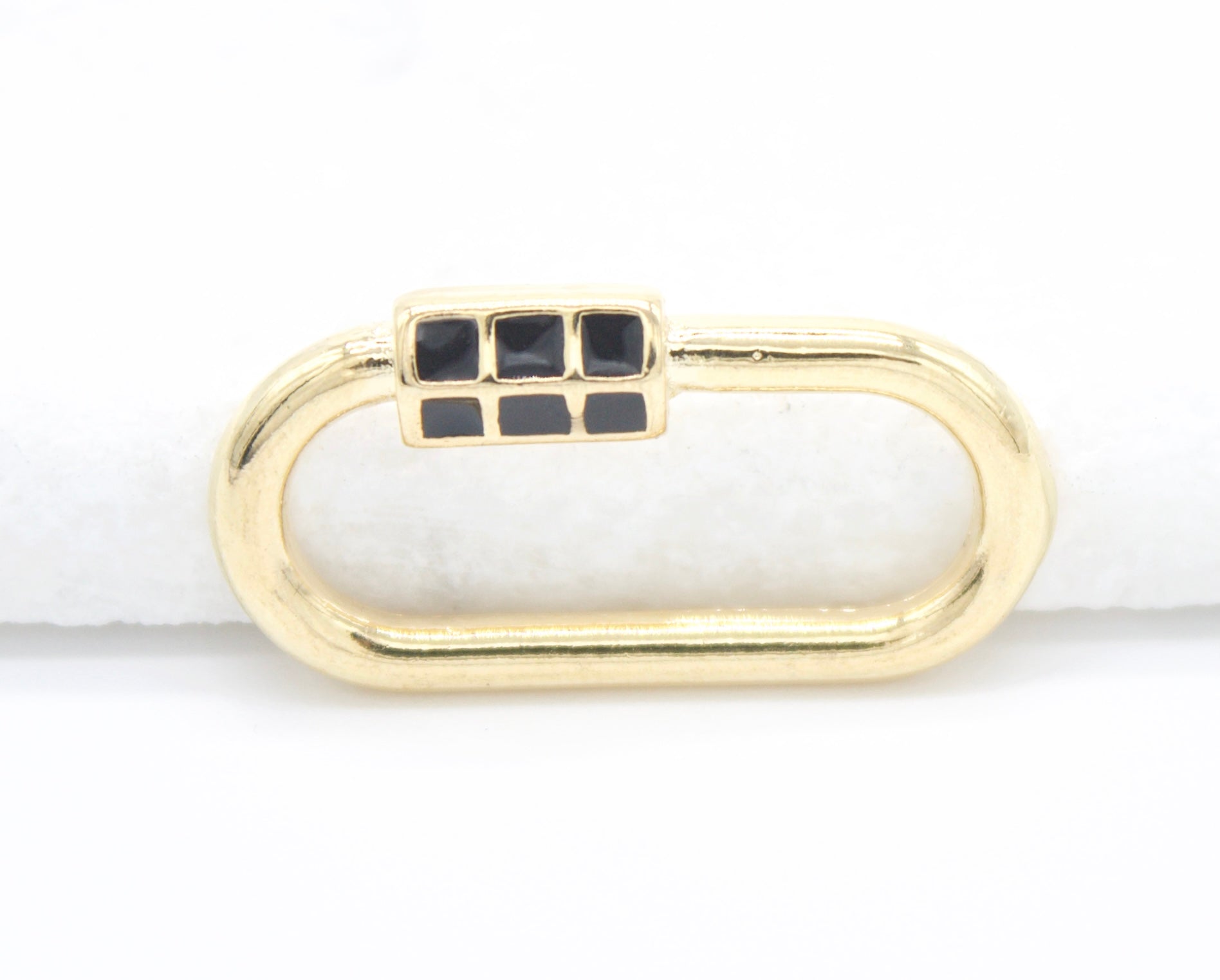 A gold oval charm connector features a black enamel grid pattern on its clasp against a white background.