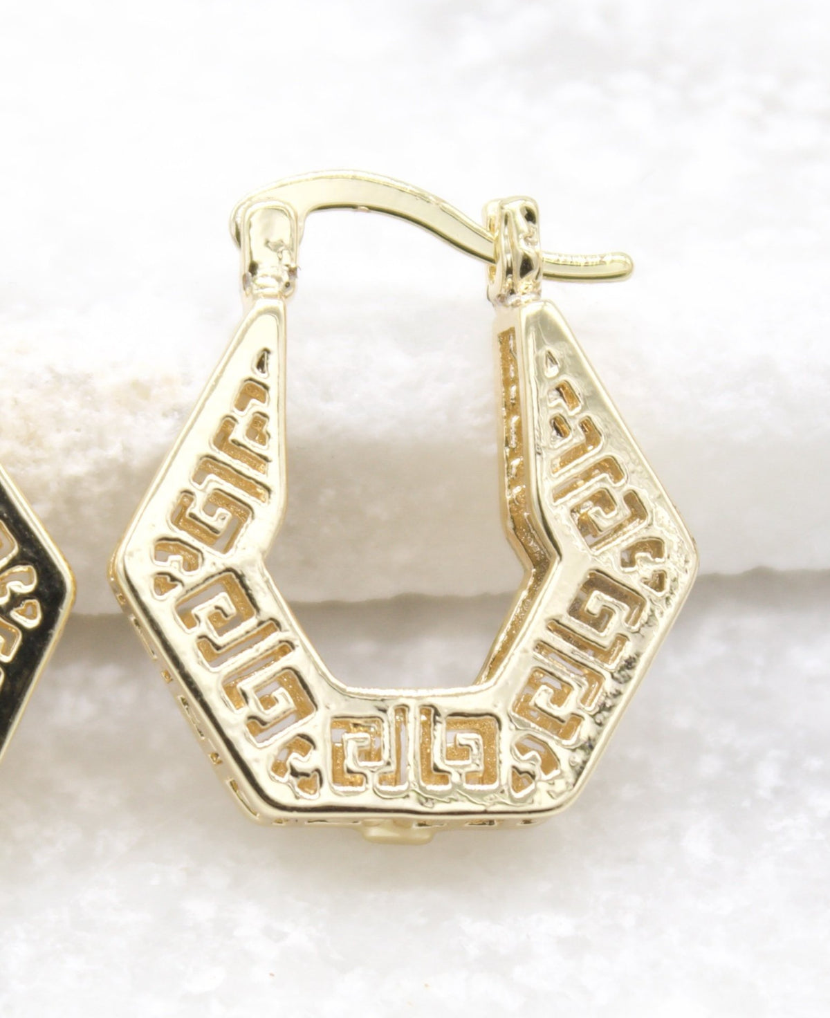 A gold hoop earring featuring an intricate geometric Greek key pattern rests against a light, textured surface.