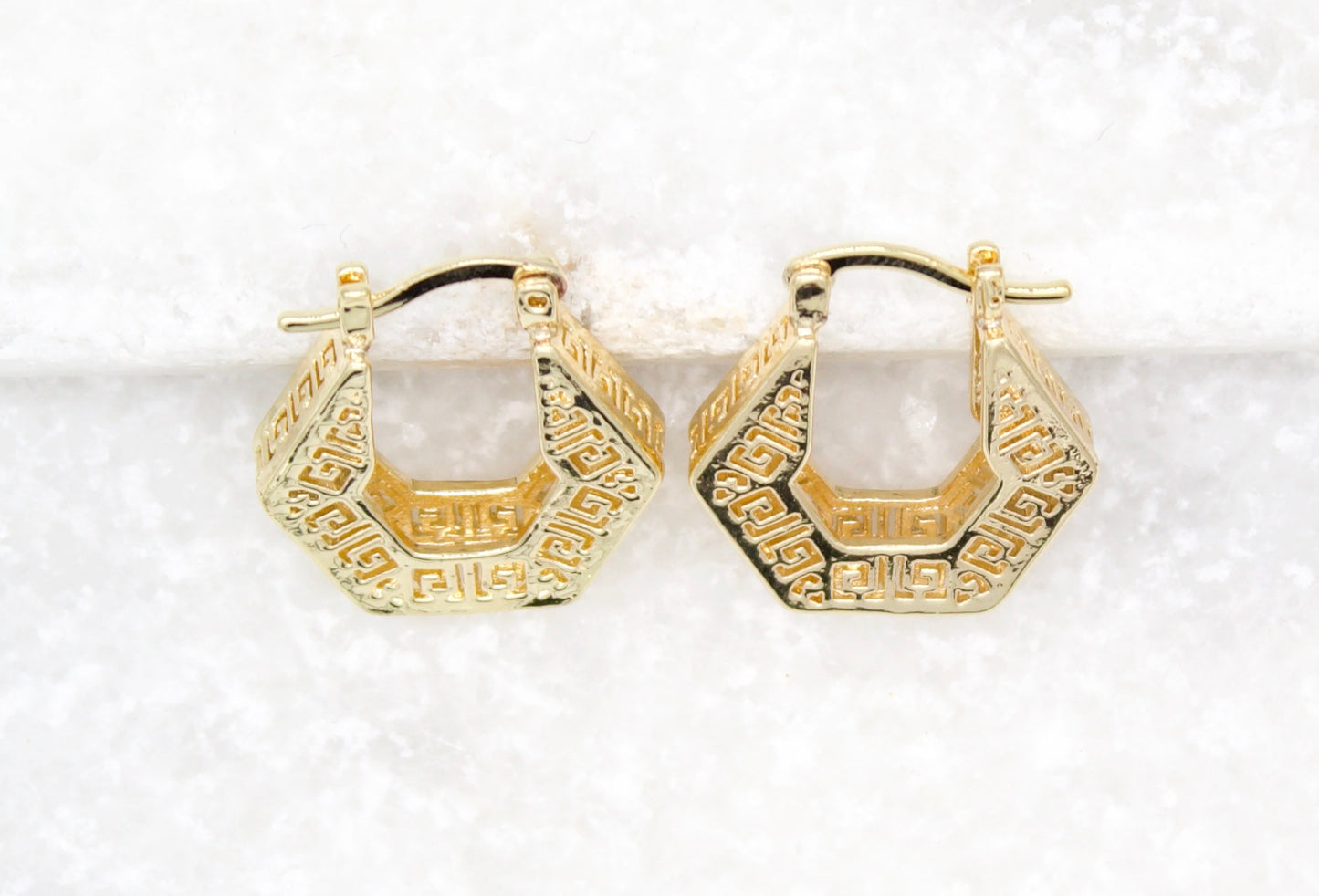 A pair of gold geometric hexagon hoop earrings featuring intricate Greek key patterns rest against a white textured background.