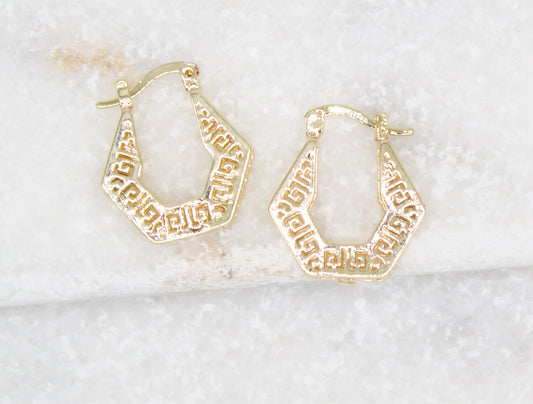 A pair of gold geometric hoop earrings rests on a white marble surface.