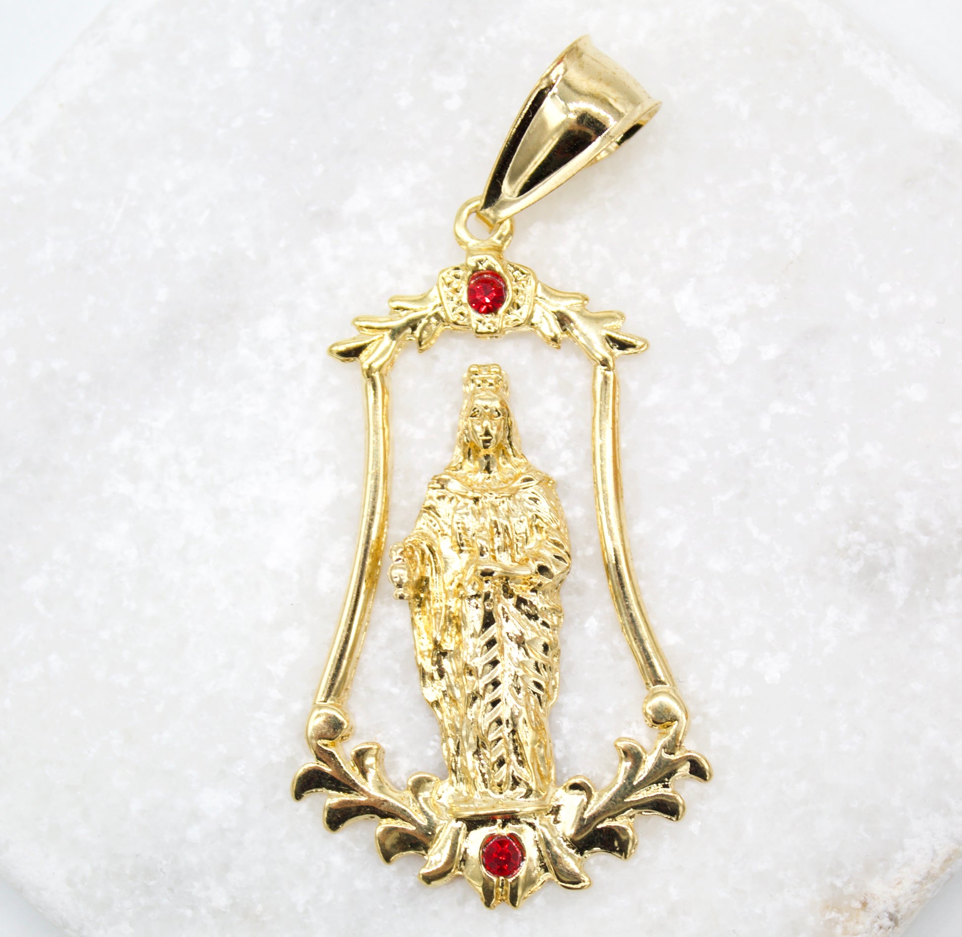 A gold pendant featuring a figure of the Virgin Mary framed by ornate scrolling details and accented with two small red stones rests on a textured white surface.