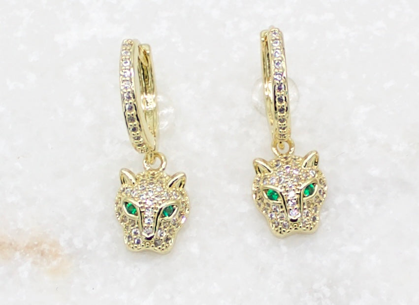A pair of gold pave-set CZ panther head huggie hoop earrings featuring vibrant green crystal eyes rests on a white background.