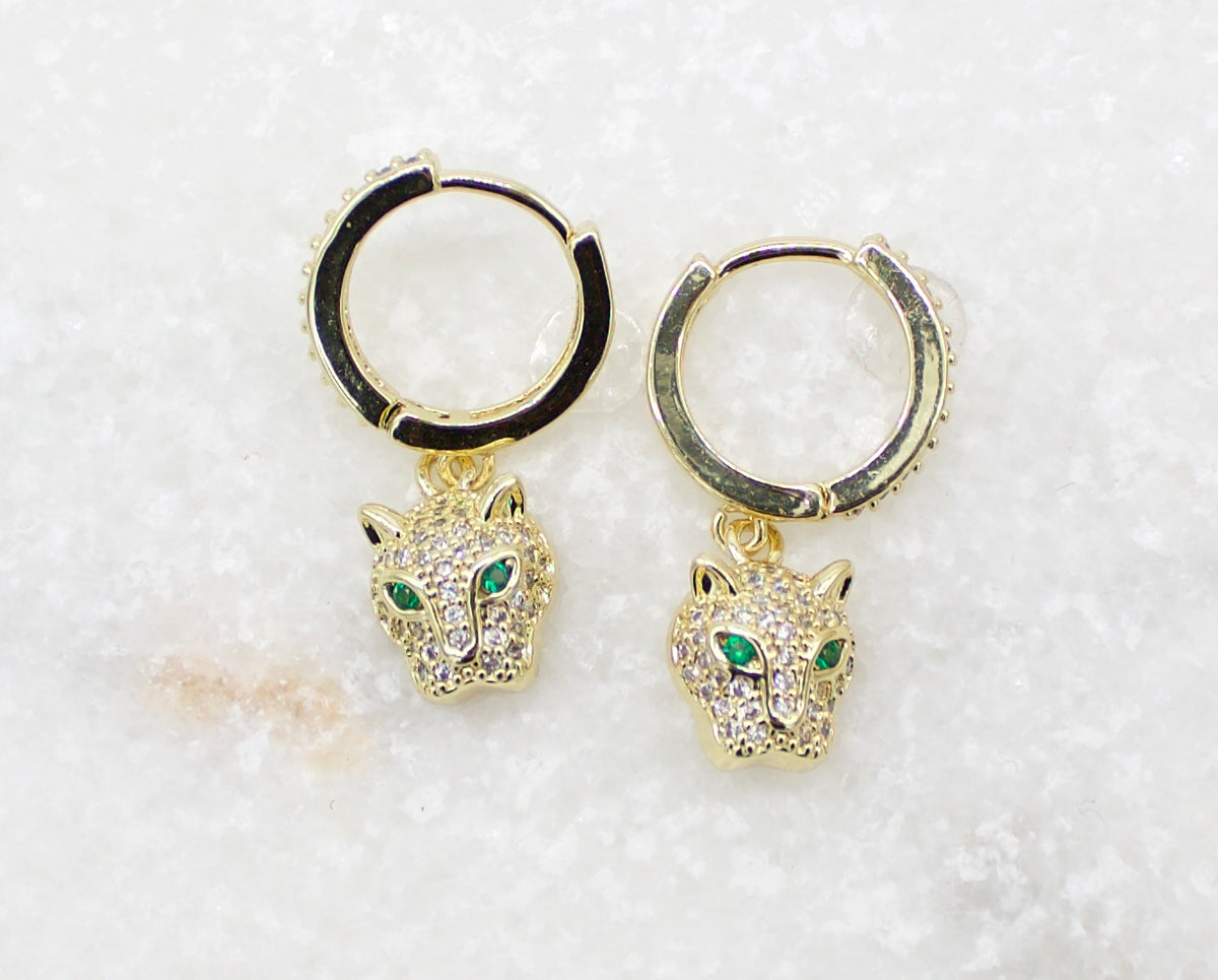 A pair of gold huggie hoop earrings featuring pavé-set crystal leopard charms with vivid green eyes rests on a white marble surface.