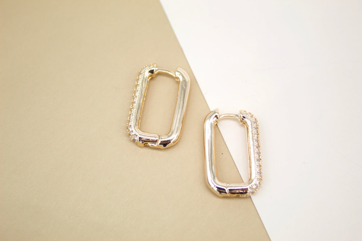A pair of elegant rectangular gold hoop earrings with sparkling pave-set stones rest on a split beige and white background.