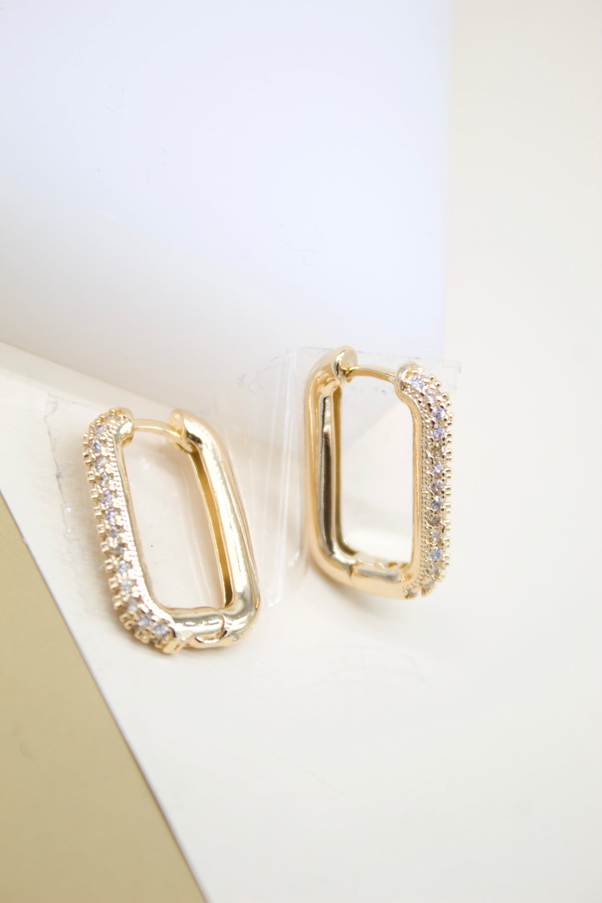 A pair of rectangular gold hoop earrings with sparkling cubic zirconia stones rest on a neutral surface.