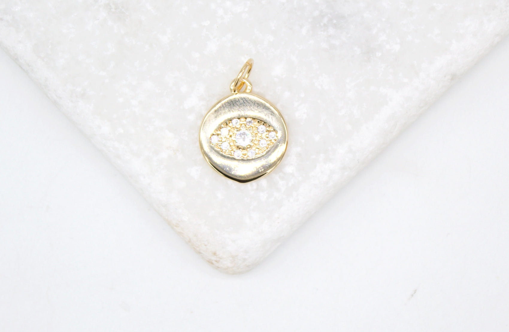 A gold evil eye charm pendant encrusted with sparkling cubic zirconia stones rests elegantly on a white marble surface.