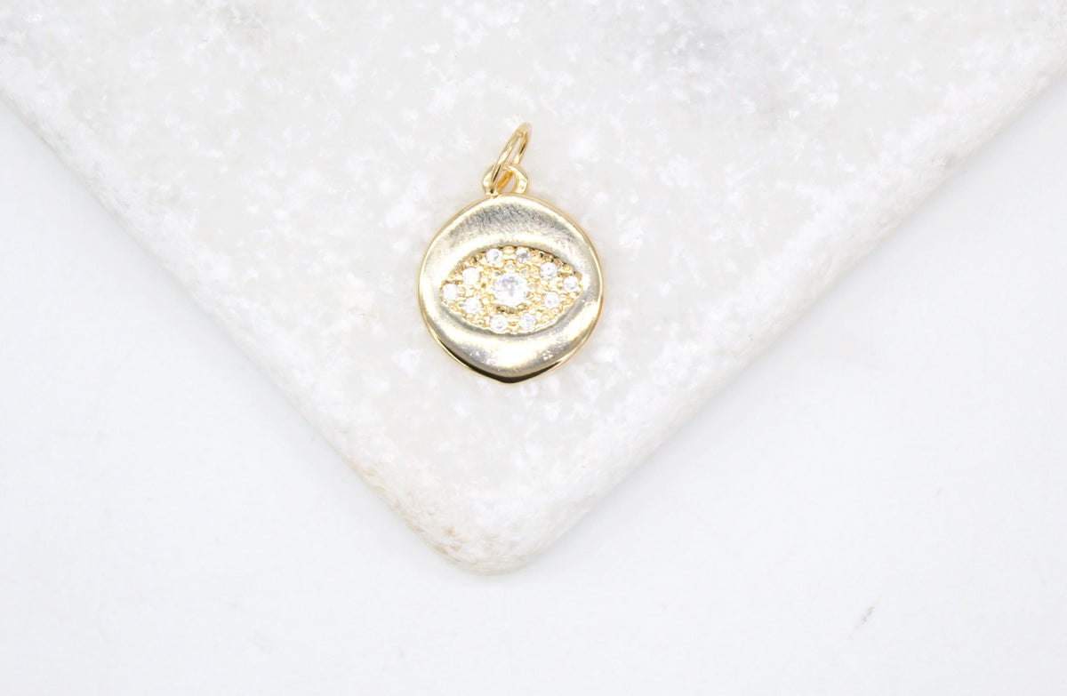A gold evil eye charm pendant encrusted with sparkling cubic zirconia stones rests elegantly on a white marble surface.