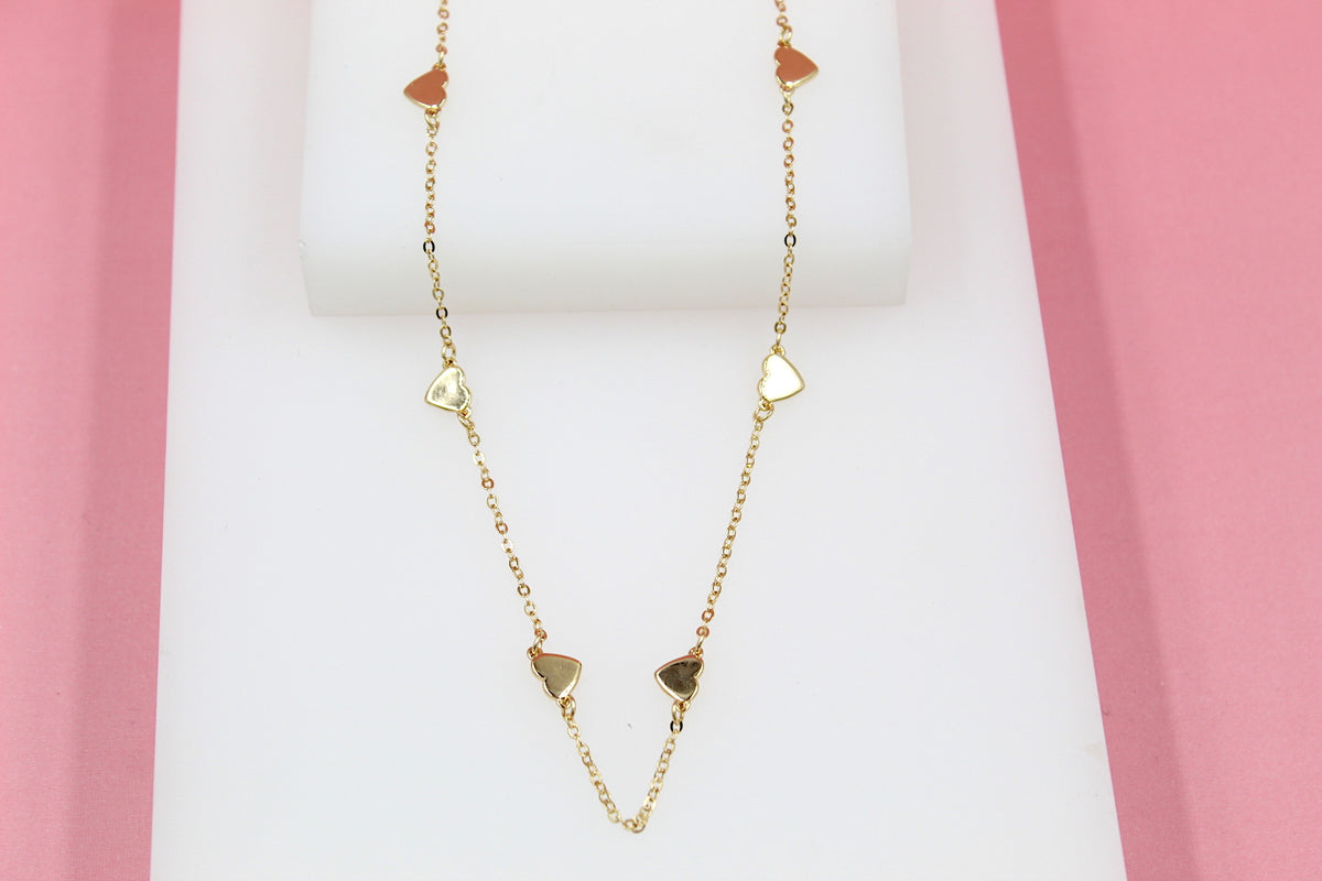A delicate gold chain necklace with interspersed heart charms rests on a white display against a pink background.