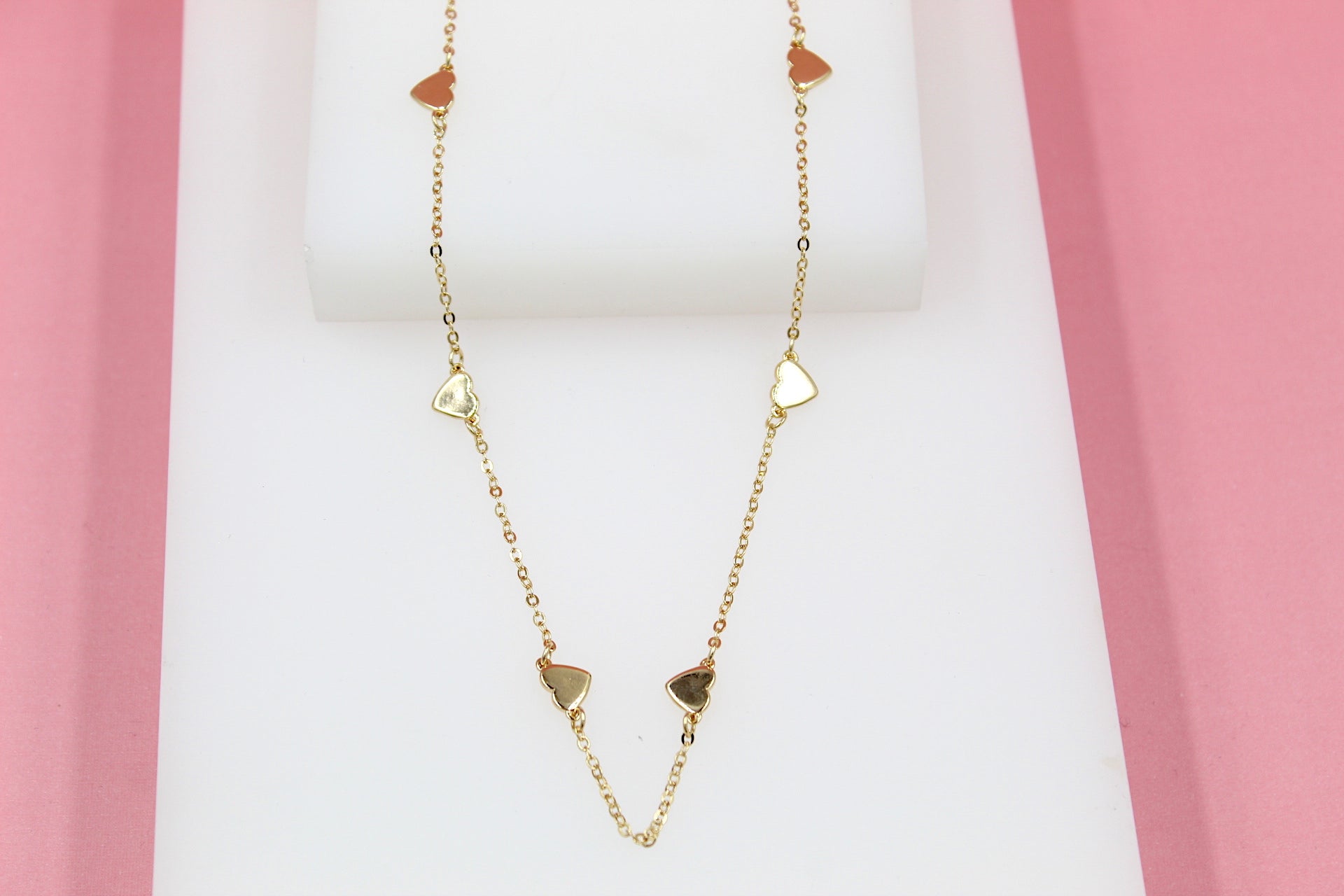 A delicate gold chain necklace with interspersed heart charms rests on a white display against a pink background.