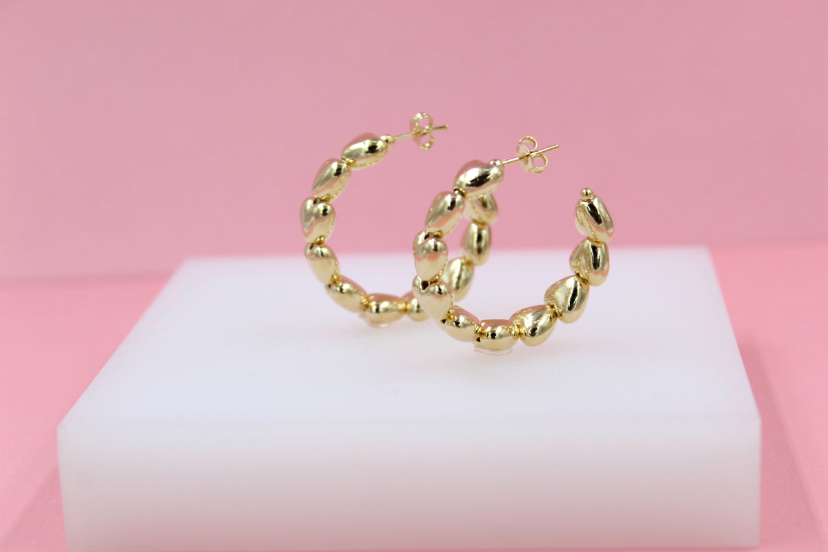 A pair of gold bubble hoop earrings rests on a white display block against a soft pink background.