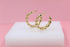 A pair of gold bubble hoop earrings rests on a white display block against a soft pink background.