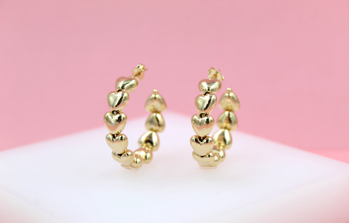A pair of polished gold heart-shaped hoop earrings rests upright on a light surface against a soft pink background.