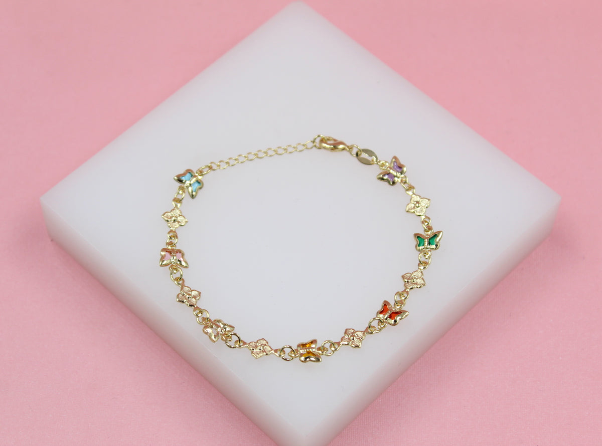 A delicate gold butterfly bracelet featuring colorful enamel accents rests on a white display block against a soft pink background.