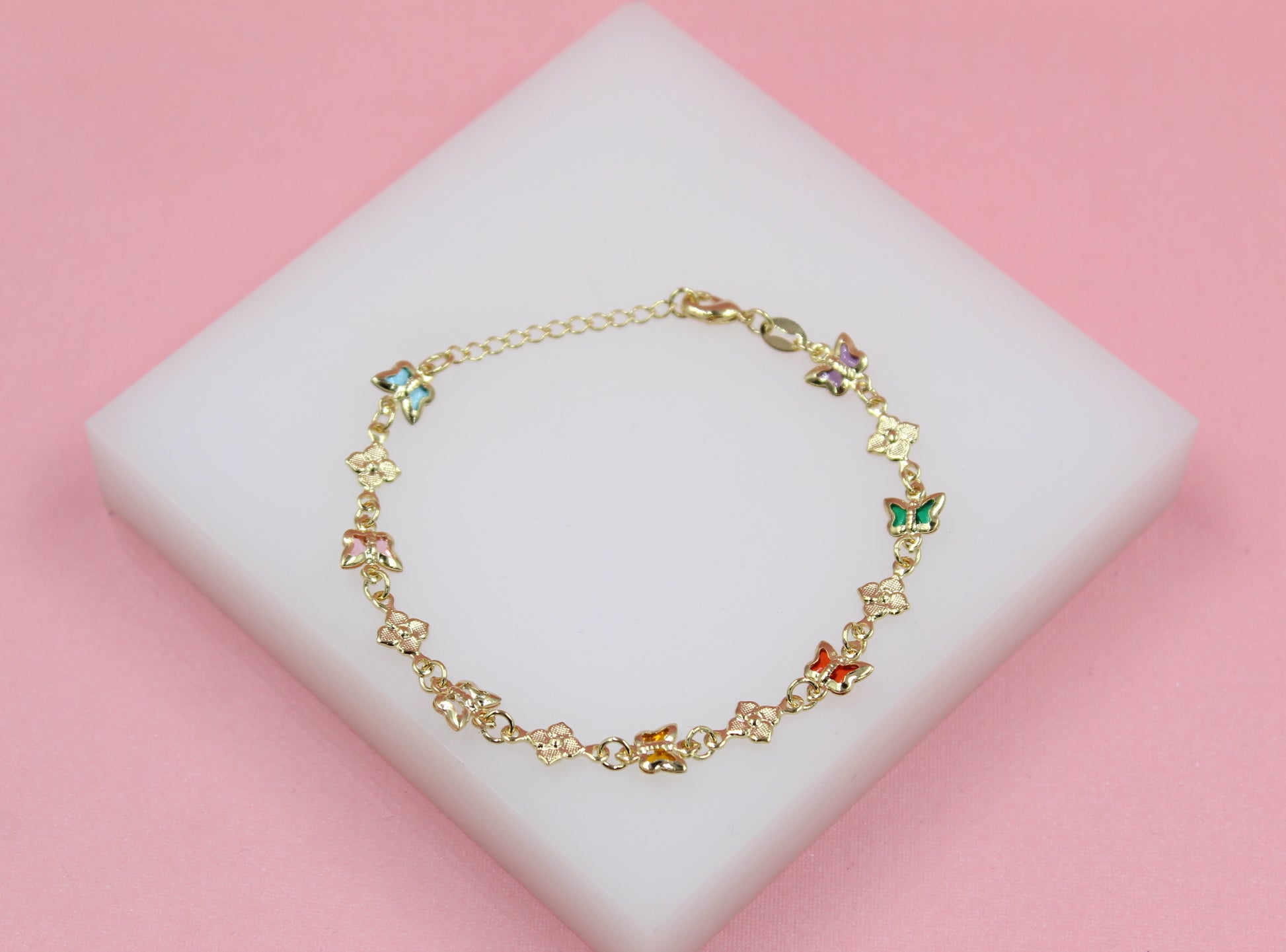 A delicate gold butterfly bracelet featuring colorful enamel accents rests on a white display block against a soft pink background.