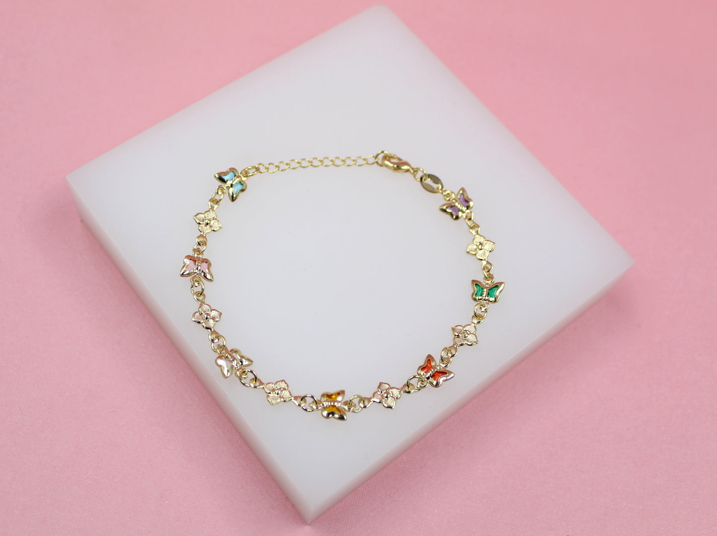 A delicate gold butterfly bracelet rests on a white display block against a pink background.