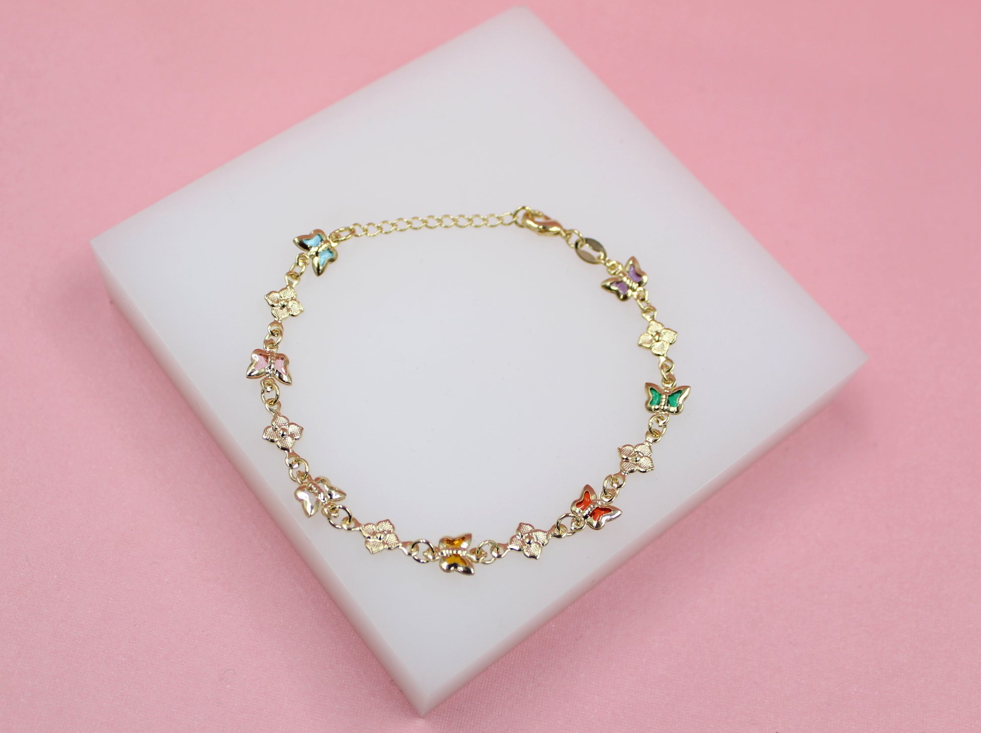 A delicate gold butterfly bracelet rests on a white display block against a pink background.