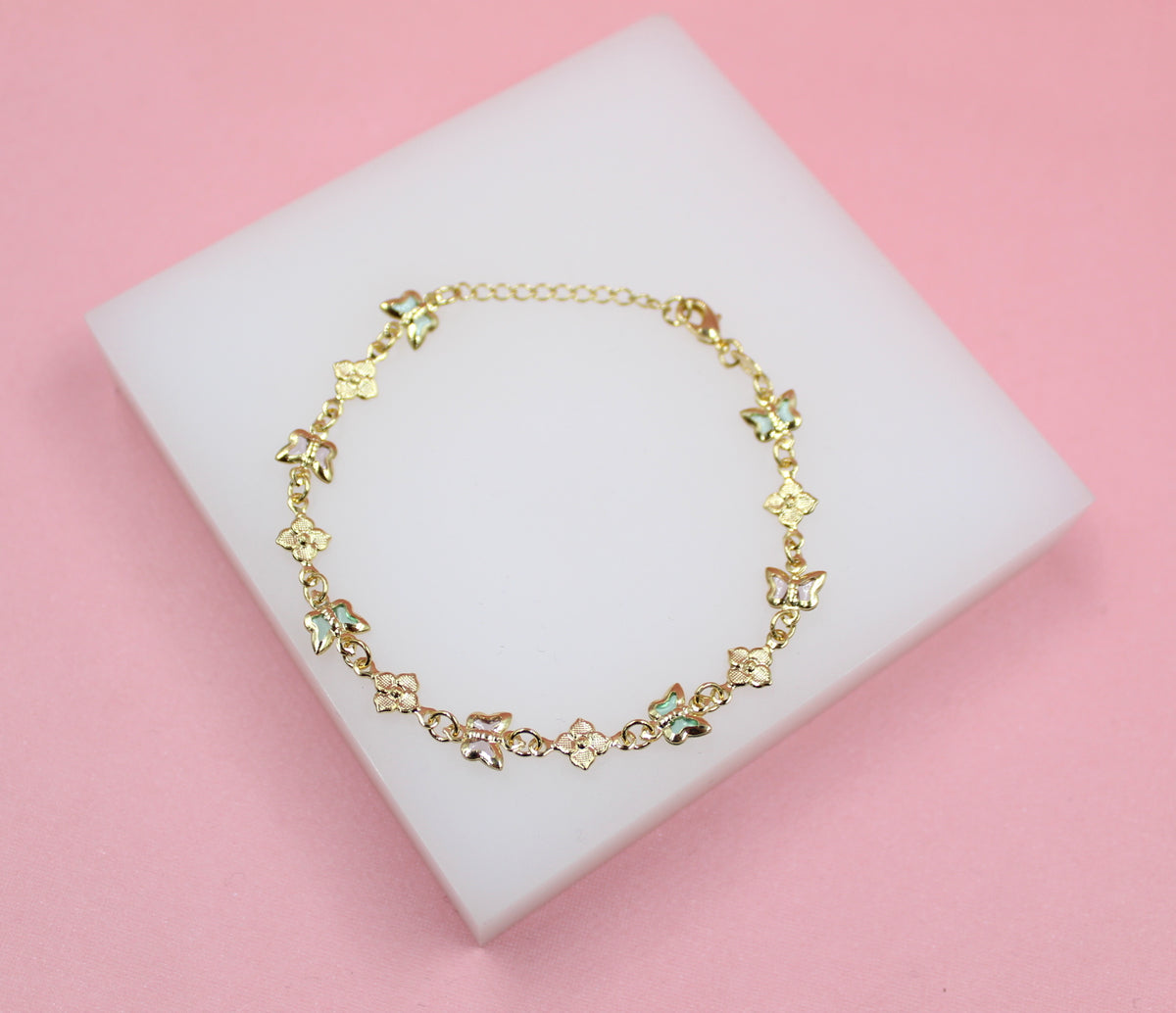 A delicate gold butterfly bracelet rests on a white display block against a pink background.