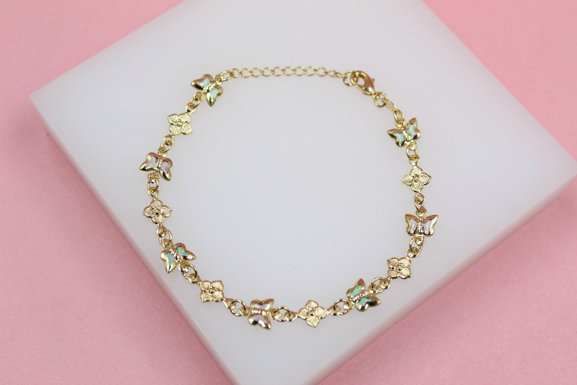 A delicate gold bracelet featuring alternating butterfly and flower charms rests on a white display block against a soft pink background.