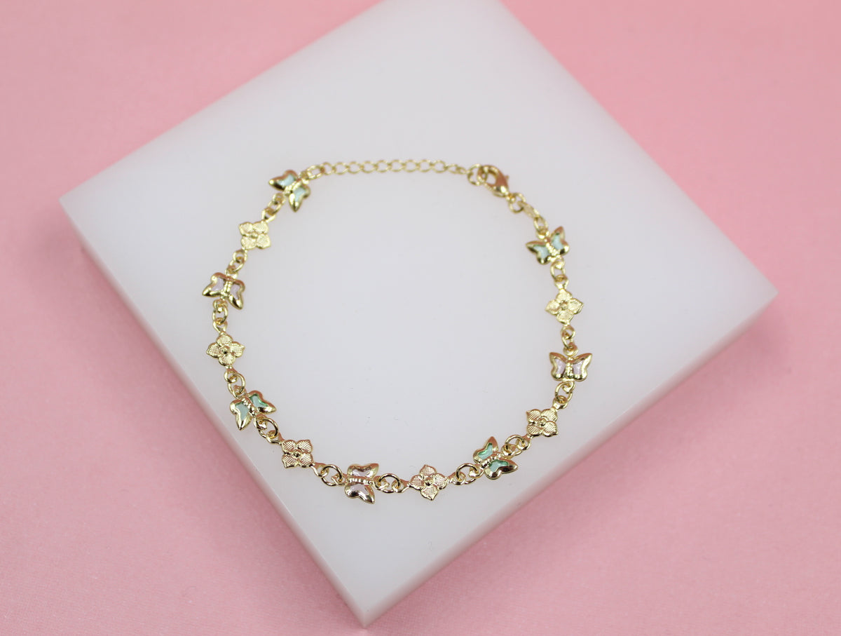 A delicate gold charm bracelet featuring alternating butterfly and flower motifs rests on a white display block against a soft pink background.