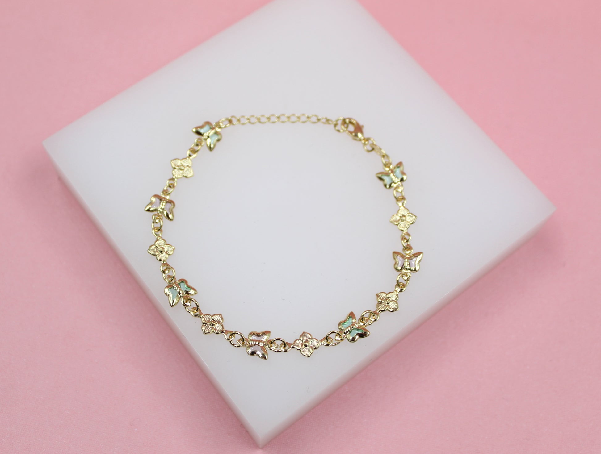 A delicate gold charm bracelet featuring alternating butterfly and flower motifs rests on a white display block against a soft pink background.