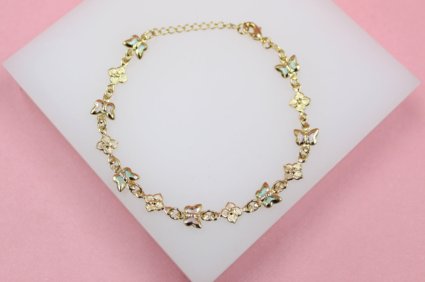 A delicate gold bracelet featuring alternating enameled butterfly charms and floral links rests on a white display surface against a pink background.
