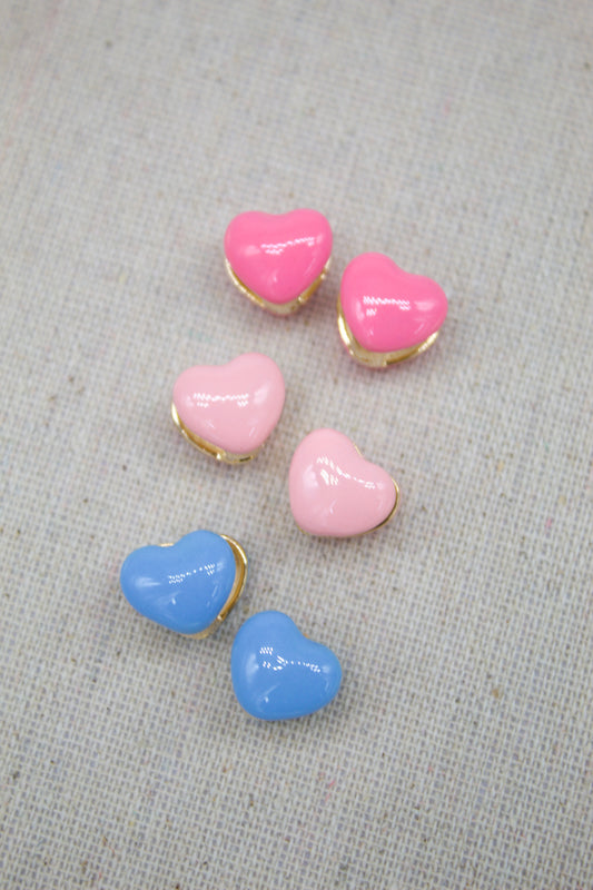 Three pairs of glossy heart-shaped huggie earrings in shades of pink, light pink, and blue rest on a light fabric surface.