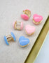 Three pairs of colorful heart-shaped huggie hoop earrings in pink, light pink, and blue rest on a textured fabric surface.