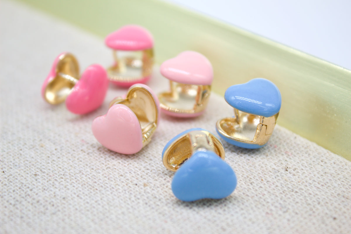 Several colorful enamel heart huggie hoop earrings in pink and blue rest on a light fabric surface.
