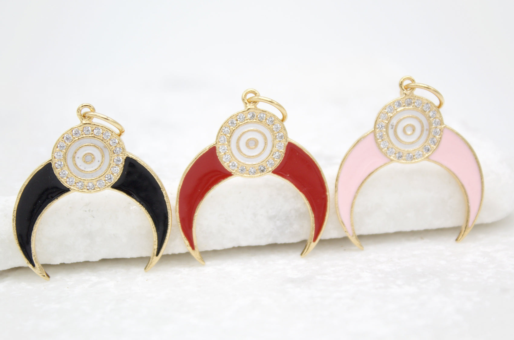 Three crescent moon evil eye pendants in black, red, and pink enamel rest on a white surface.