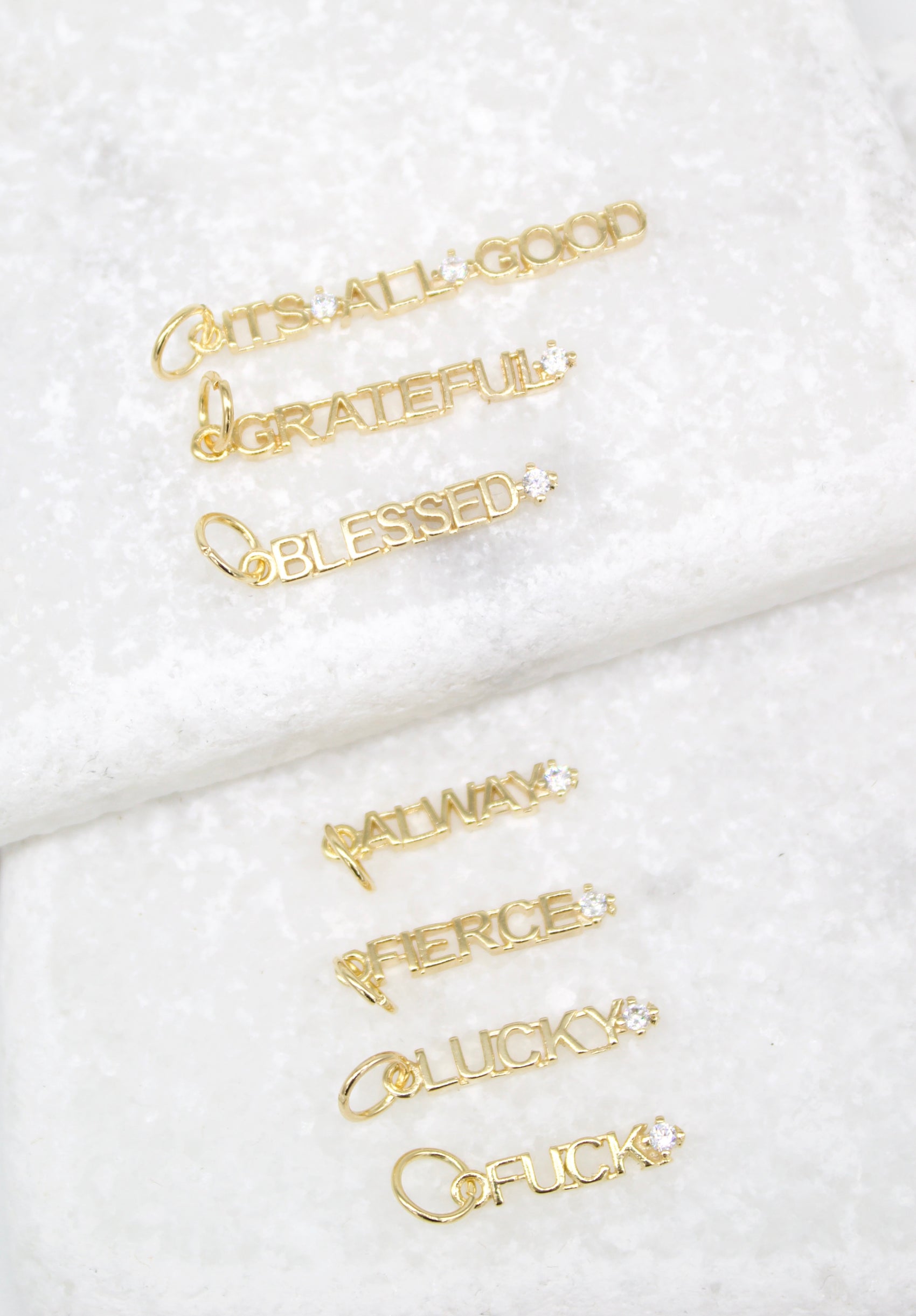 Several gold word charms featuring motivational inscriptions and sparkling diamond accents lie arranged on a white marble surface.