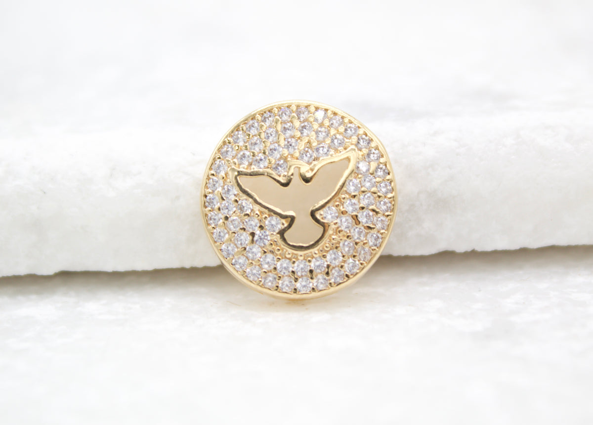 A gold pendant rests on a white surface, featuring a pavé-set crystal background framing a polished silhouette of a flying dove.