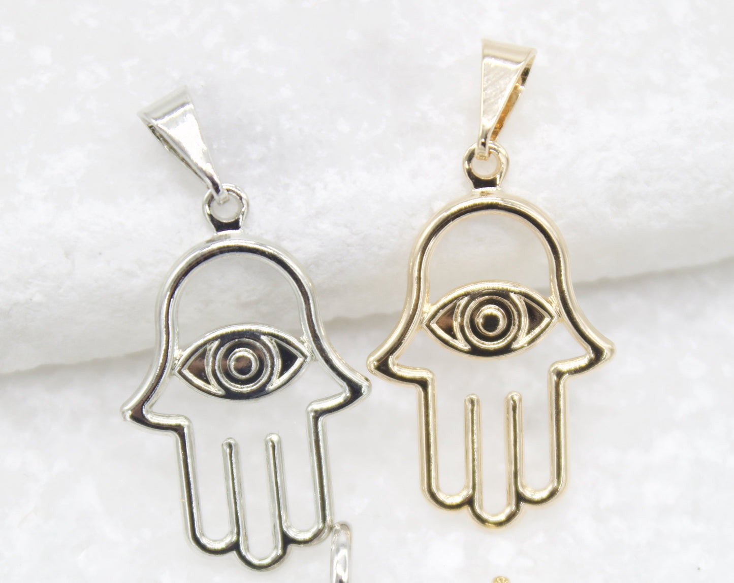 Two Hamsa hand pendants featuring evil eye designs rest side by side on a white surface, with one made of silver and the other of gold.