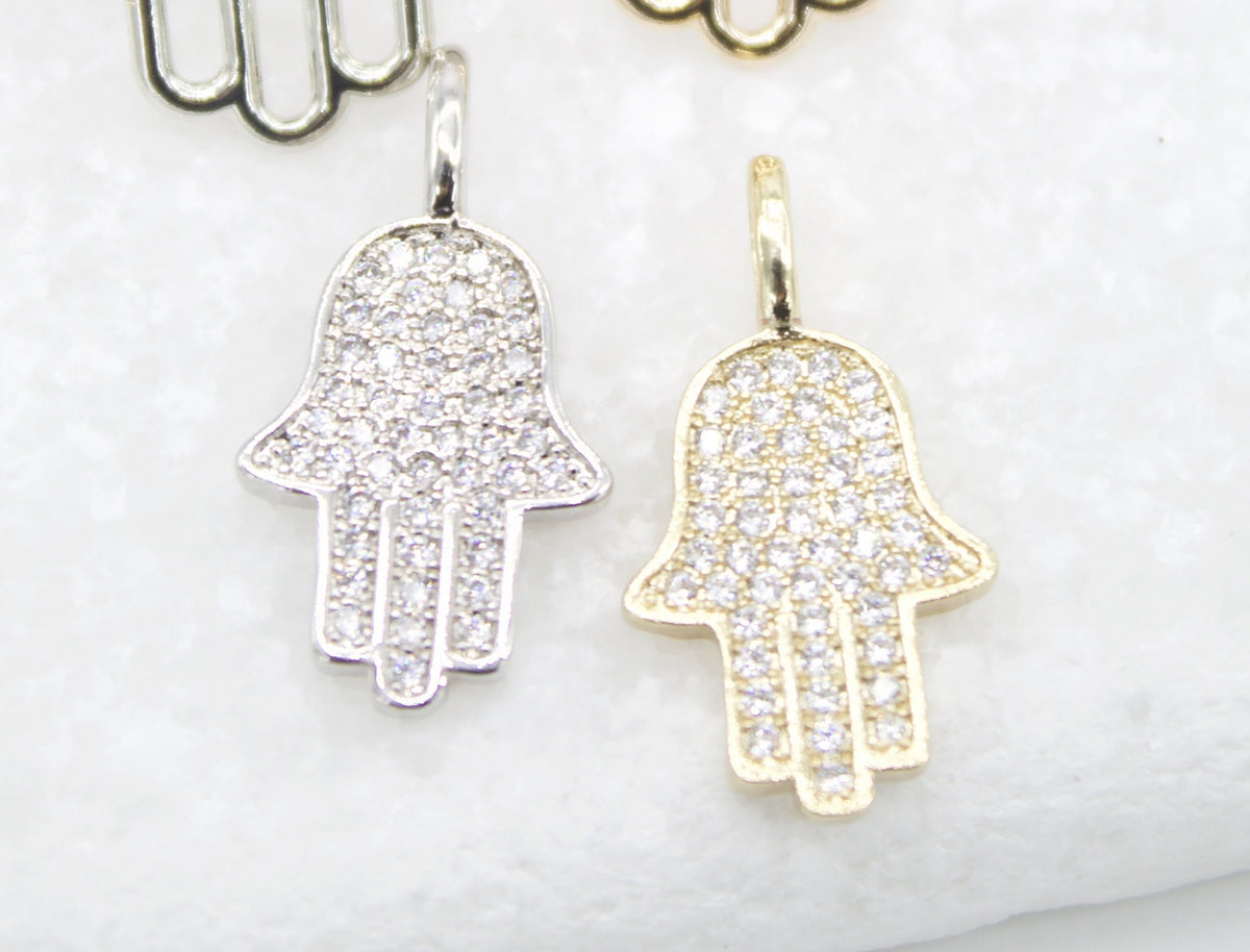 Two pavé diamond-encrusted Hamsa hand charms, one silver and one gold, rest side by side on a textured white surface.