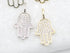 Two pavé diamond-encrusted Hamsa hand charms, one silver and one gold, rest side by side on a textured white surface.