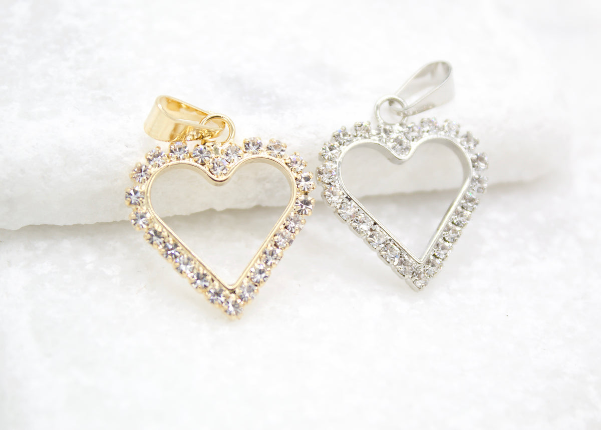 Two sparkling rhinestone-encrusted heart pendants, one in gold and one in silver, rest side-by-side on a textured white surface.