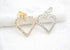 Two sparkling rhinestone-encrusted heart pendants, one in gold and one in silver, rest side-by-side on a textured white surface.