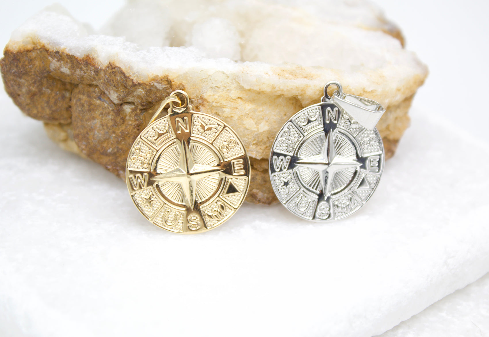 A gold compass rose pendant and a silver compass rose pendant rest side by side on a crystal geode.
