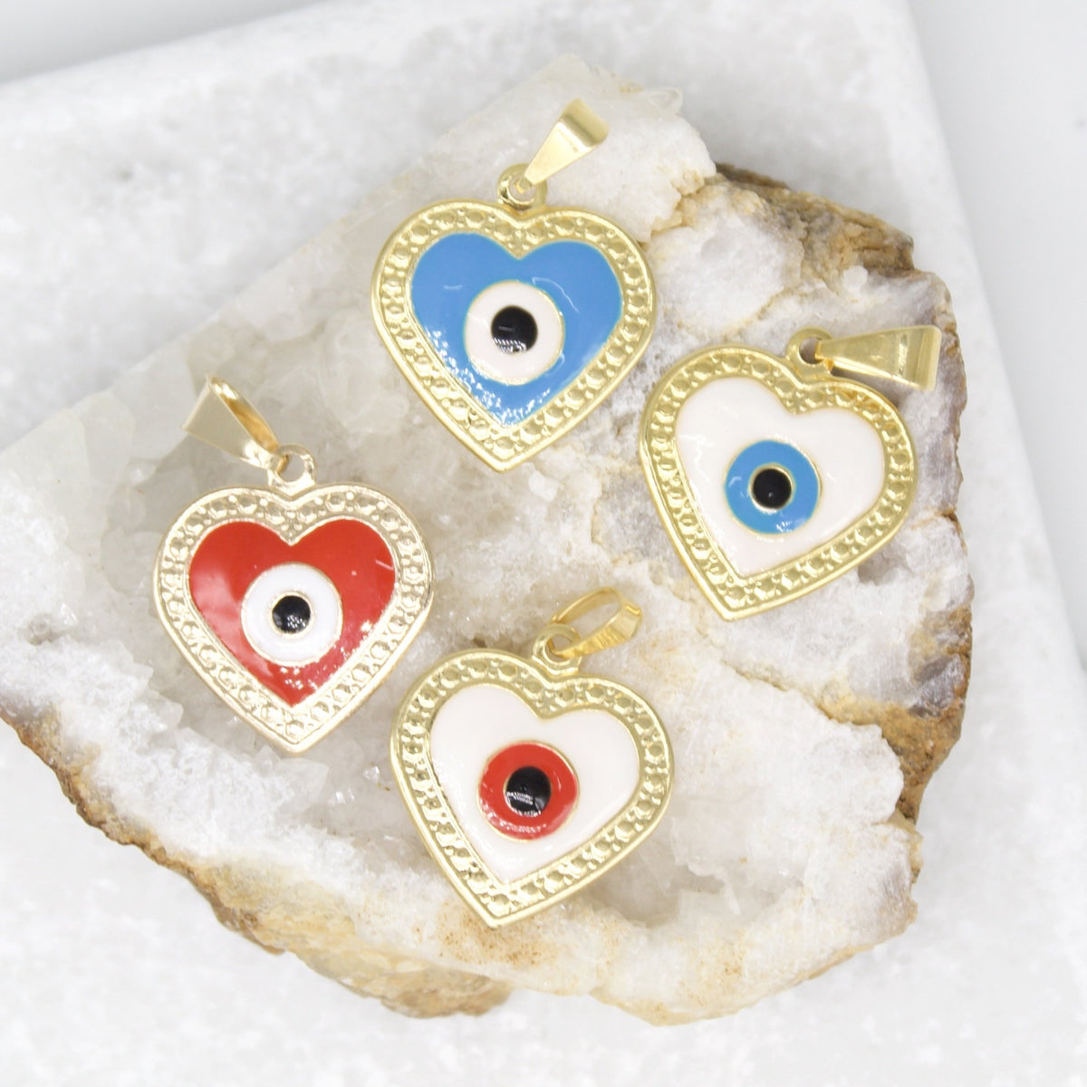 Four gold evil eye heart pendants in red, blue, and white enamel rest delicately on a sparkling white quartz crystal cluster.