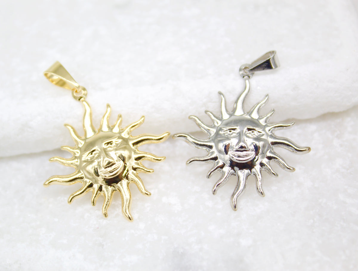 Two shiny celestial sun face pendants resting side-by-side on a textured white surface display contrasting gold and silver finishes.