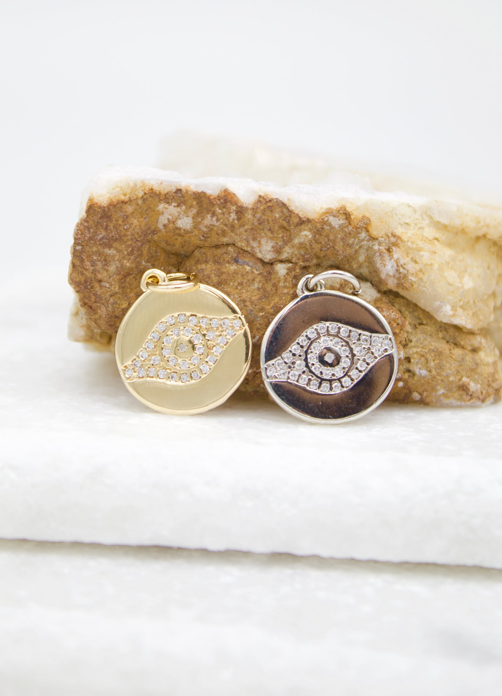 Two round evil eye pendants, one gold and one silver with a black enamel background, rest side-by-side on a rustic stone surface.