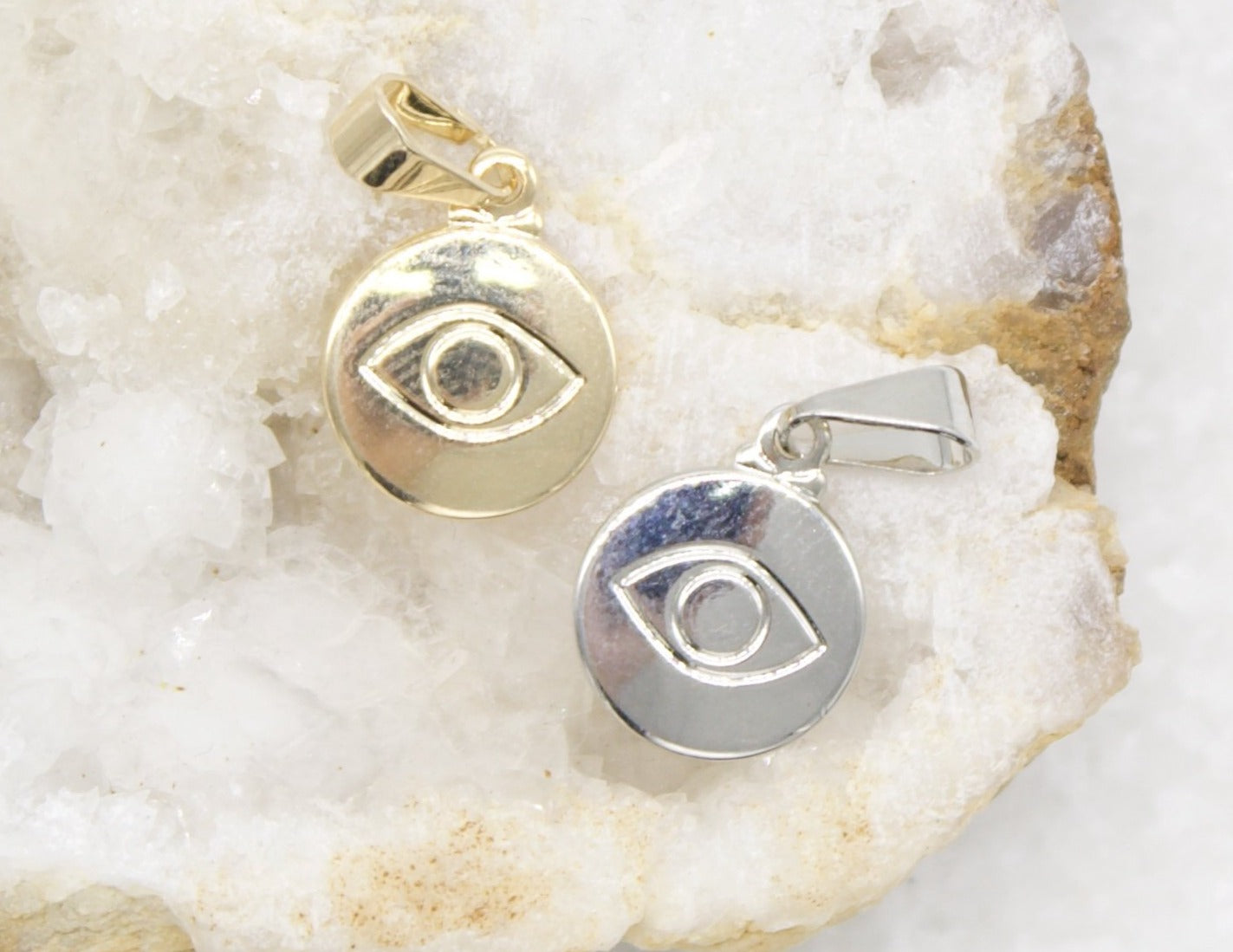 A gold evil eye pendant and a silver evil eye pendant rest side-by-side on a textured crystal surface.