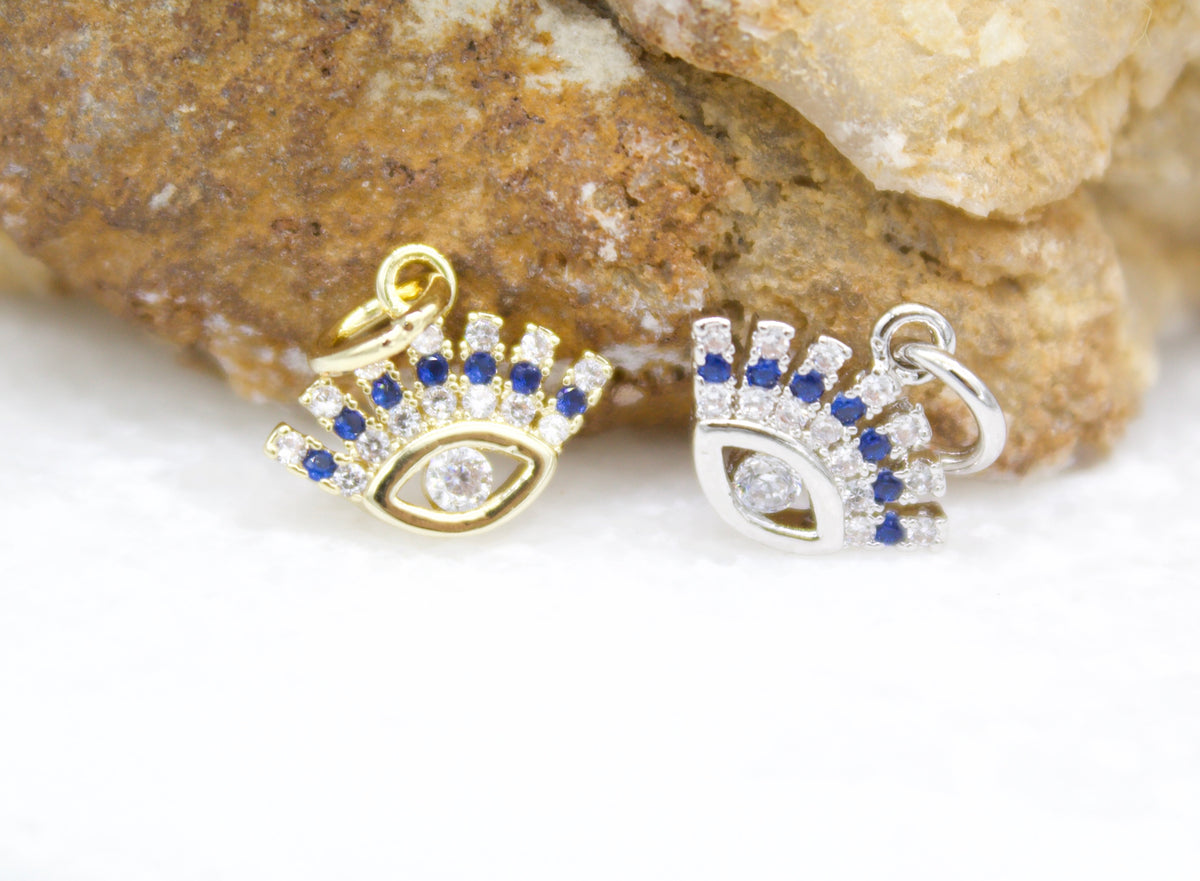 A gold and a silver evil eye charm, each embedded with sparkling white and blue cubic zirconia, rest against a textured stone backdrop.
