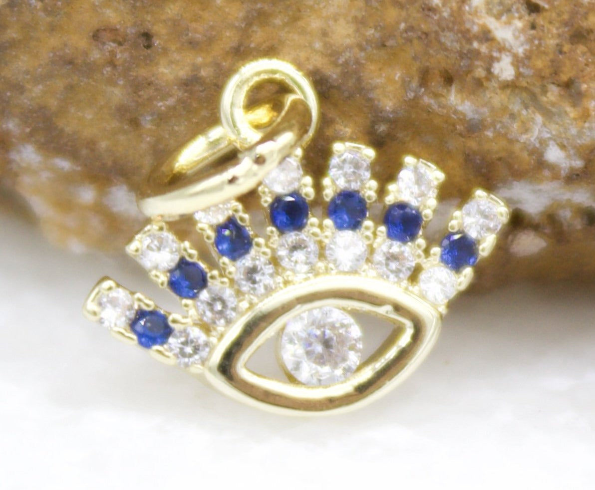 A gold evil eye charm sparkles with embedded white diamonds and blue sapphires against a textured rock background.