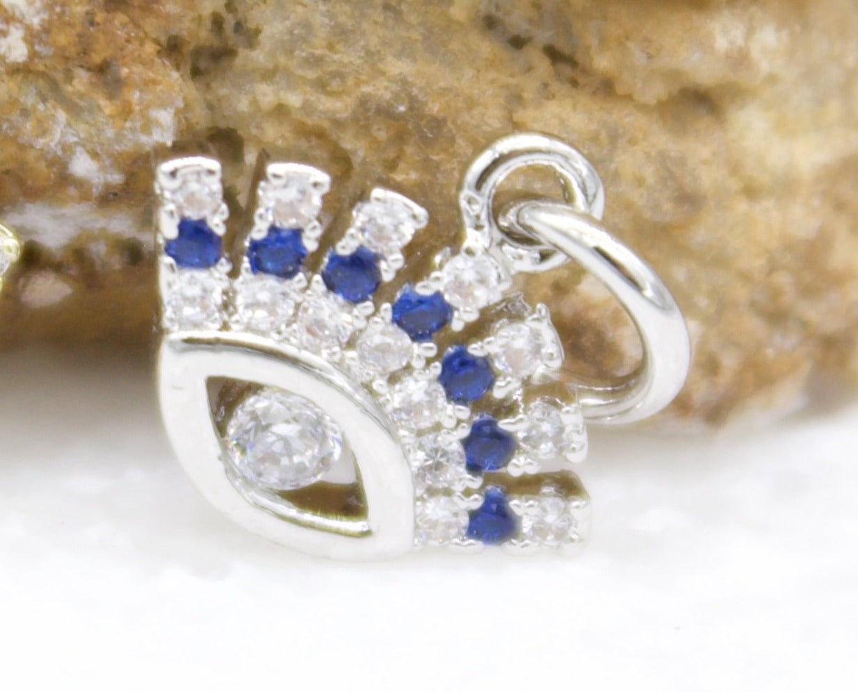 A silver evil eye charm sparkles with pave-set white and blue cubic zirconia stones against a textured stone backdrop.