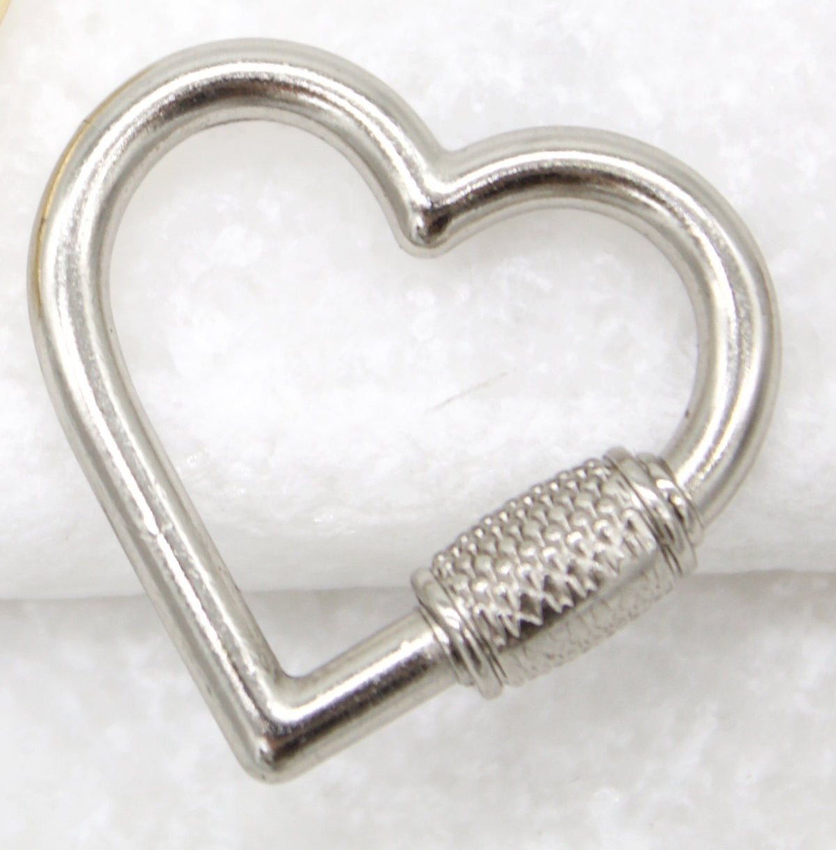 A shiny silver heart-shaped carabiner rests on a textured white surface.